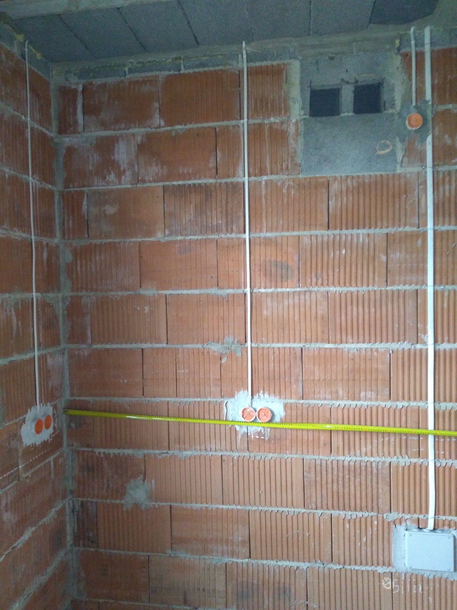 Ceglana ściana w trakcie prac elektrycznych: białe kable prowadzone pionowo, pomarańczowe puszki instalacyjne i żółta miarka pozioma. Surowy stan wykończenia.
