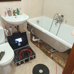 AQUAPATROL ROBERT KORNECKI - Inspekcja pod wanną z użyciem kamery inspekcyjnej w kufrze, widoczne przewody i elementy mocujące wannę, uszkodzona podłoga w łazience, w tle umywalka i toaleta.