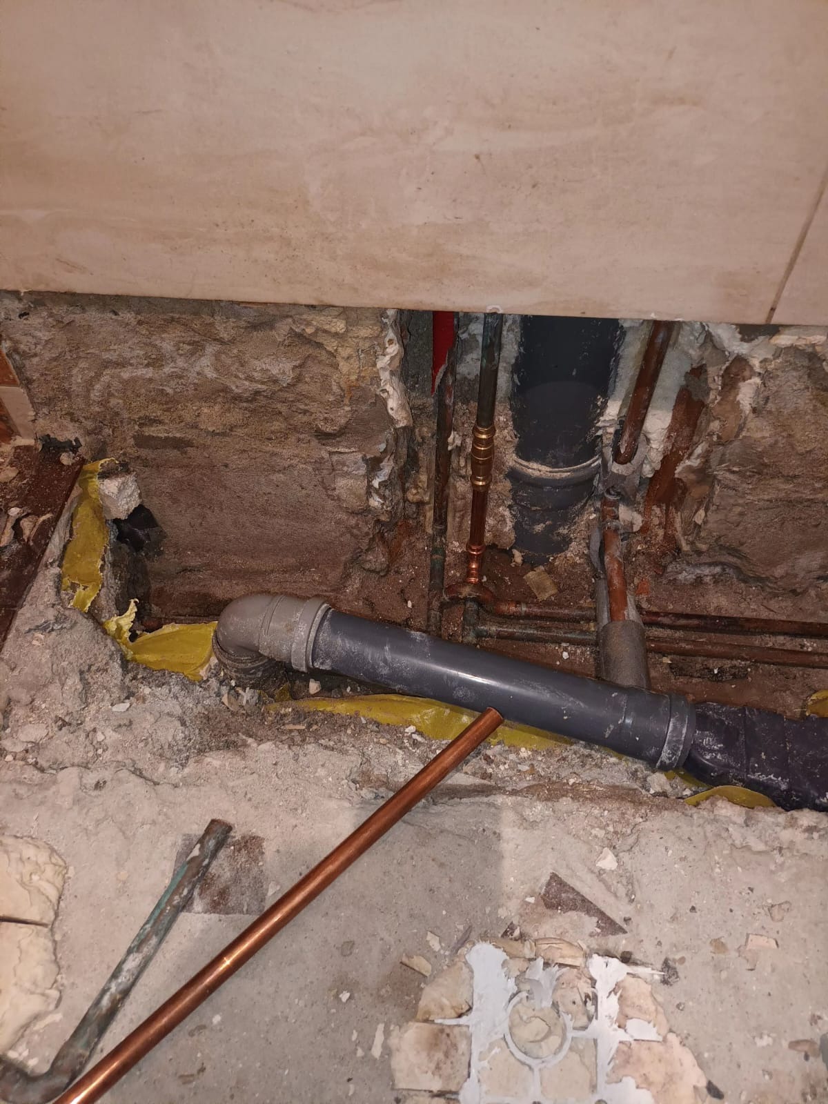 Odkryte rury kanalizacyjne i miedziane w ścianie podczas remontu, widoczna żółta izolacja i gruz.