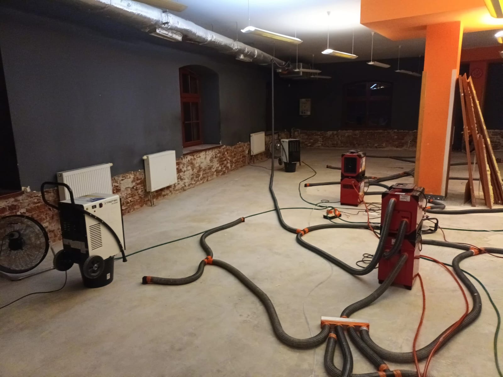 Wnętrze w trakcie osuszania: czerwone i białe osuszacze podłączone do elastycznych przewodów ułożonych na surowej podłodze, ściany z widoczną cegłą, wentylacja pod sufitem.