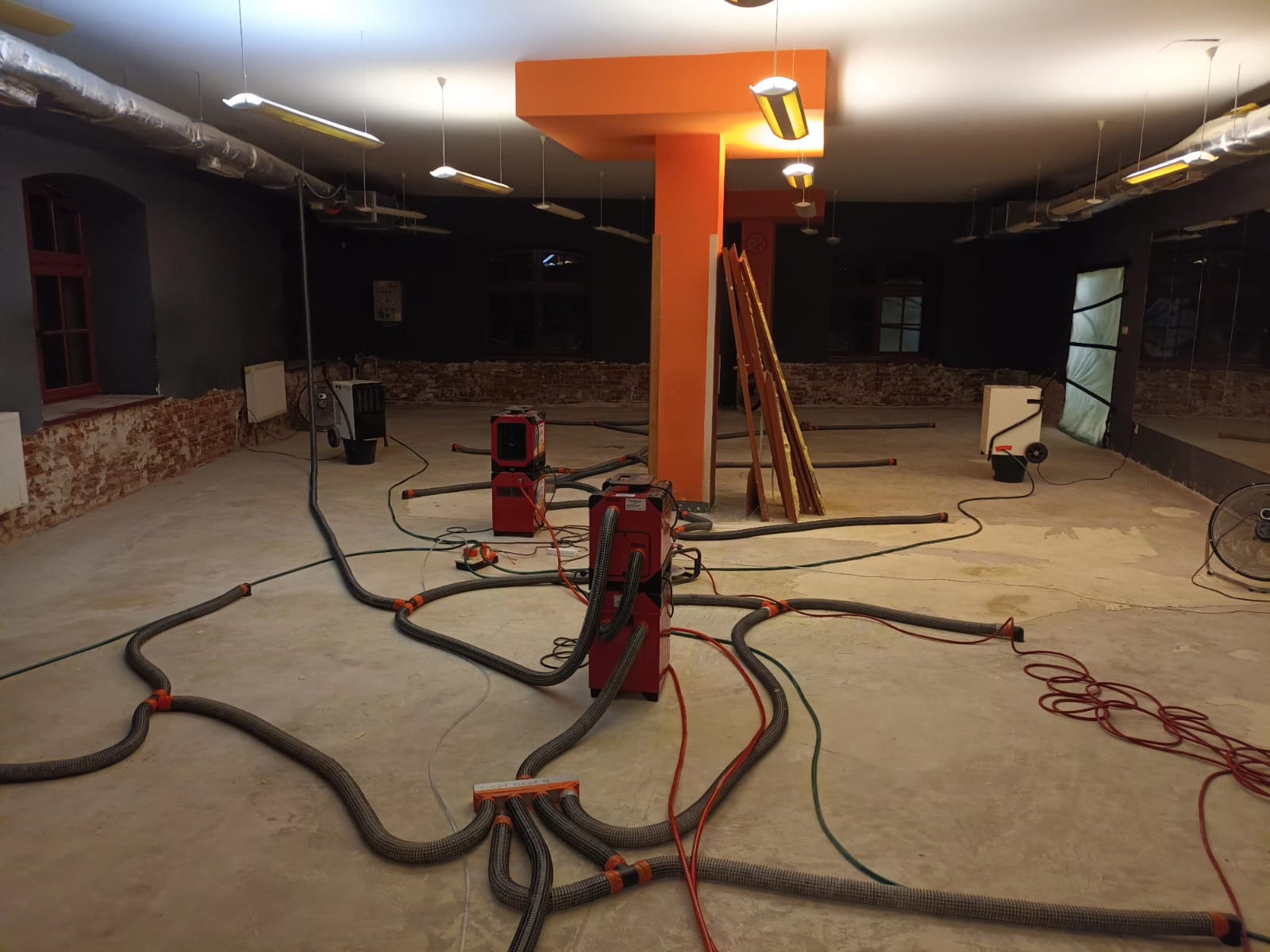 Wnętrze pomieszczenia w trakcie osuszania, widoczne czerwone urządzenia osuszające podłączone do systemu rur na posadzce, z odsłoniętą cegłą na ścianach i pomarańczowym filarem.