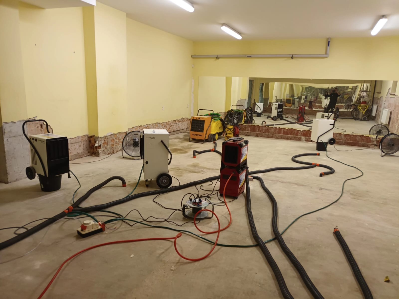 Wnętrze w trakcie osuszania z użyciem kilku urządzeń osuszających różnych marek i modeli, widoczne węże i kable rozłożone na podłodze, ściany częściowo przygotowane do remontu, lustro na ścianie...