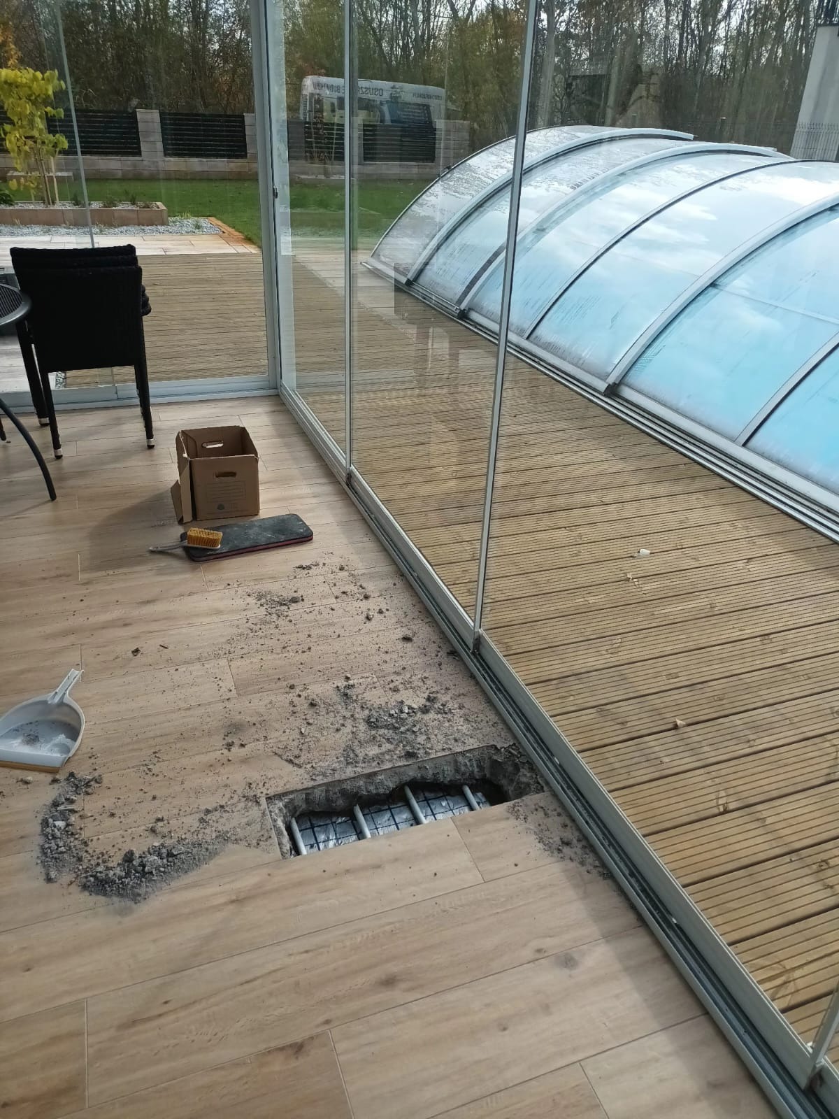 Otwór w drewnianej podłodze wewnątrz przeszklonego pomieszczenia, prawdopodobnie przygotowany pod instalację osuszacza; widoczne zbrojenie i resztki gruzu, obok leży zmiotka i szufelka.