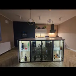 Nowoczesna kuchnia z wyspą wyposażoną w przeszkloną szafkę ekspozycyjną na alkohole, oświetloną od wewnątrz, na podłodze parkiet w jodełkę.