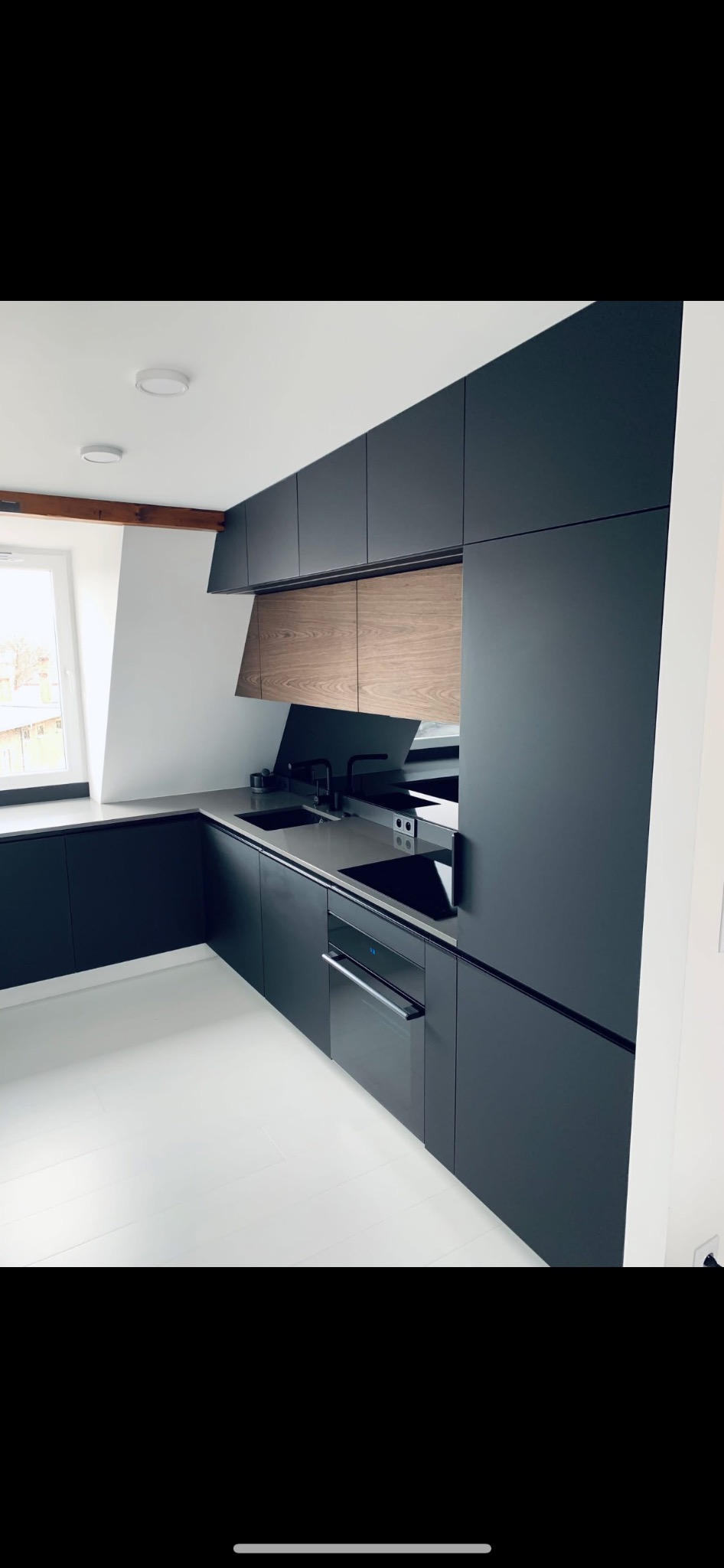 Nowoczesna kuchnia z ciemnymi, matowymi szafkami, drewnianymi akcentami nad blatem, czarną armaturą i jasną podłogą, oświetlona światłem dziennym z okna.