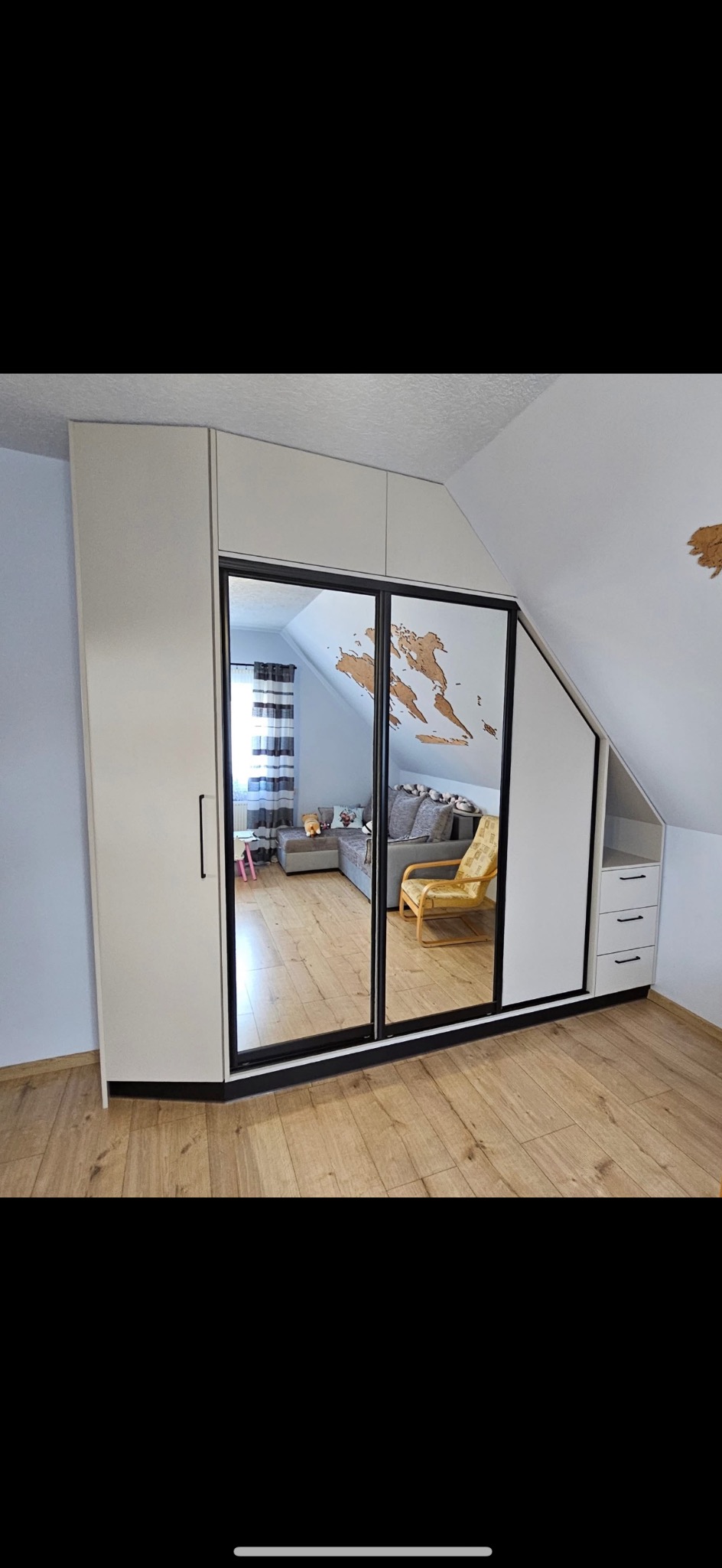 Biała szafa wnękowa z lustrzanymi drzwiami przesuwnymi, widoczne odbicie pokoju z kanapą i żółtym fotelem, poddasze ze skośnym sufitem i drewnianą mapą świata na ścianie.