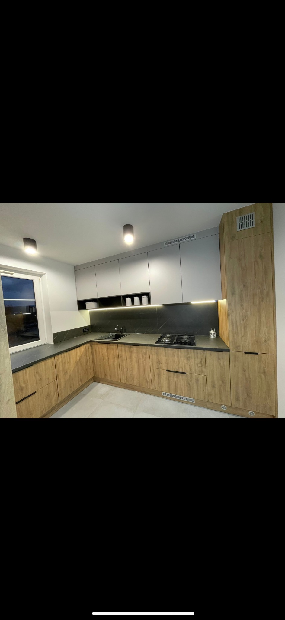 Nowoczesna kuchnia w kształcie litery L z drewnianymi frontami szafek dolnych i szarymi górnymi, czarny blat i podświetlenie LED pod szafkami wiszącymi, okno z widokiem na zewnątrz.