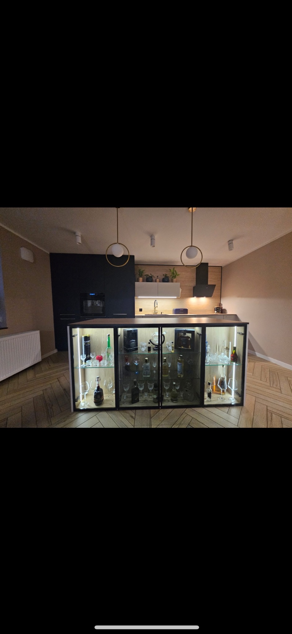 Nowoczesna kuchnia z wyspą wyposażoną w przeszkloną szafkę ekspozycyjną na alkohole, oświetloną od wewnątrz, na podłodze parkiet w jodełkę.