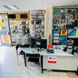 Wnętrze serwisu komputerowego z ladą, komputerem stacjonarnym, akcesoriami komputerowymi na ścianie i otwartymi drzwiami wejściowymi.