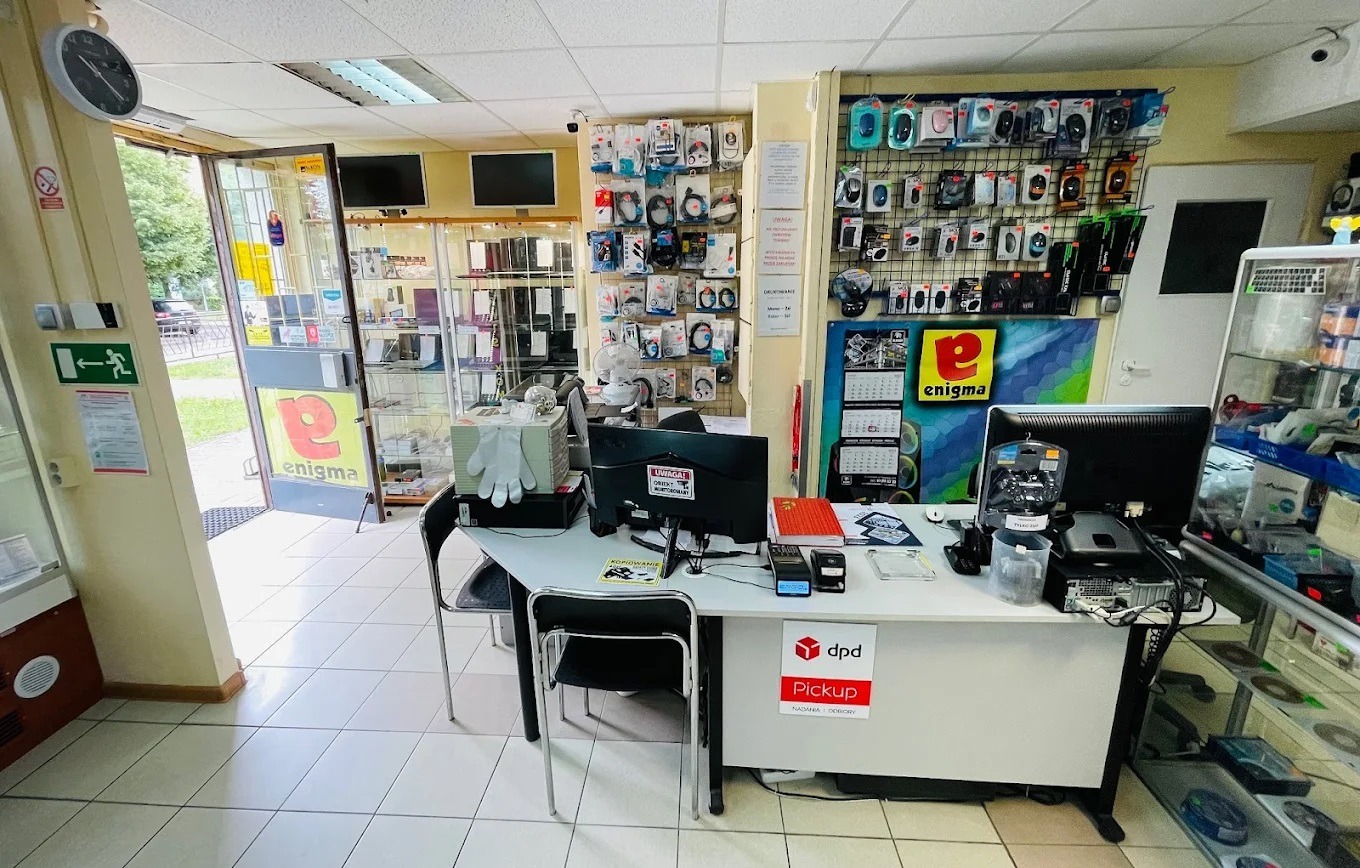 Wnętrze serwisu komputerowego z ladą, komputerem stacjonarnym, akcesoriami komputerowymi na ścianie i otwartymi drzwiami wejściowymi.