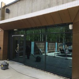 MSS MAREK WIĄCEK - Czyszczenie elewacji budynku biurowego z elementami z blachy trapezowej. Pracownik w trakcie pracy z odkurzaczem przemysłowym. Widoczne duże okna.