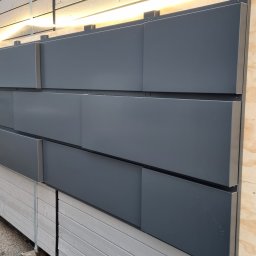MSS MAREK WIĄCEK - Nowoczesna elewacja z szarych paneli kompozytowych, podświetlona listwą LED, na tle budynku. Widoczne elementy montażowe i drewniana konstrukcja nośna.