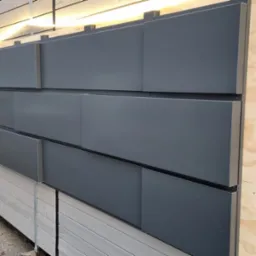 Nowoczesna elewacja z szarych paneli kompozytowych, podświetlona listwą LED, na tle budynku. Widoczne elementy montażowe i drewniana konstrukcja nośna.