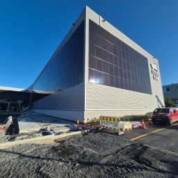 MSS MAREK WIĄCEK - Nowoczesny budynek z panelami słonecznymi na elewacji, w trakcie prac wykończeniowych terenu. Widoczny sprzęt budowlany i elementy infrastruktury.