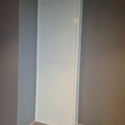 Pionowa, biała konstrukcja z paneli na tle szarej ściany i drewnianej podłogi. Widoczne łączenia paneli, oświetlenie sufitowe. Kątowe ujęcie z dołu.