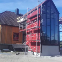 Dom w trakcie budowy z rusztowaniem, widoczne elementy konstrukcyjne i okna. Czerwona fasada kontrastuje z tradycyjną architekturą obok.