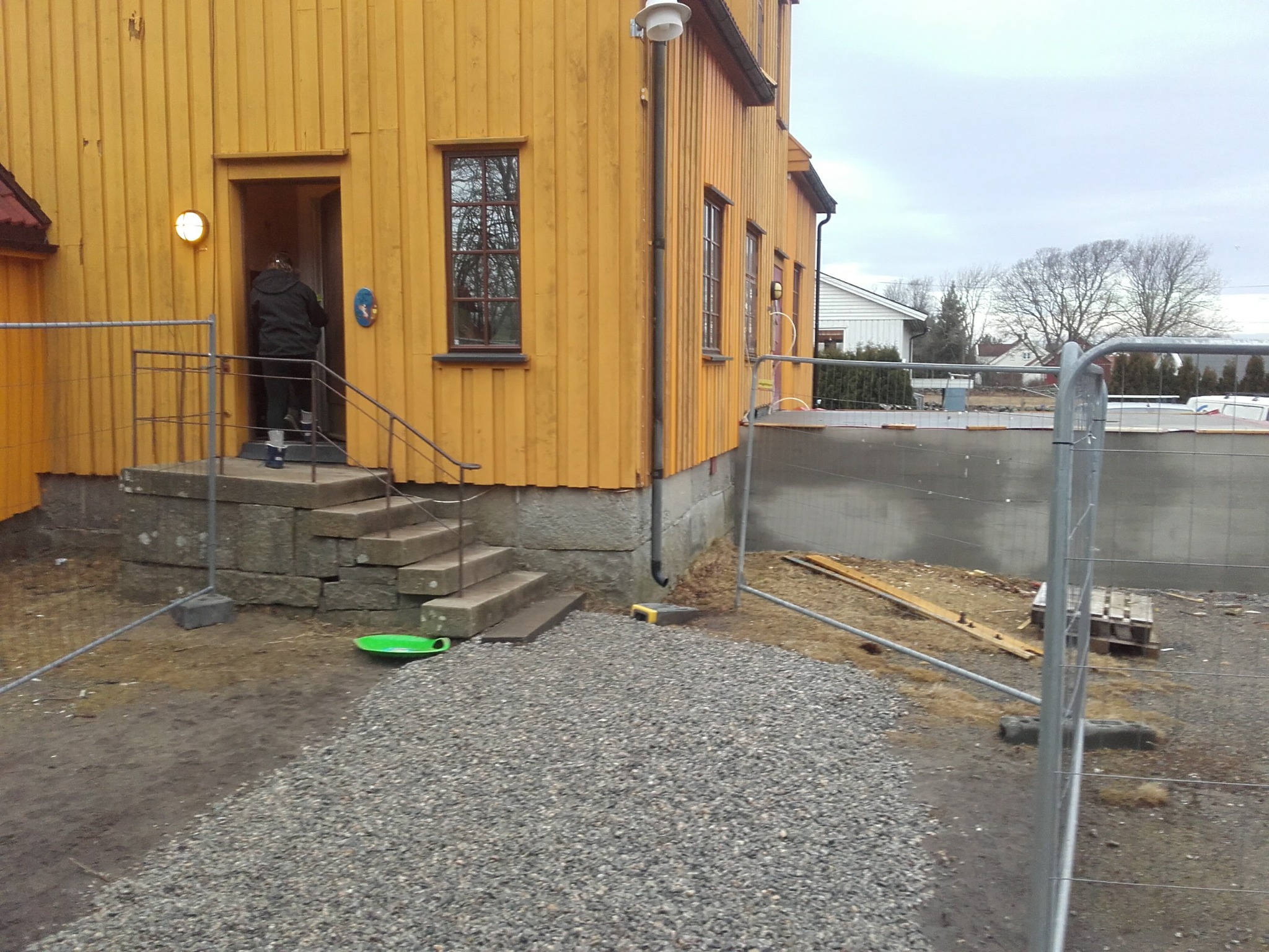 Żółty dom z drewnianą elewacją i schodami wejściowymi. Widoczna osoba wchodząca do budynku i teren budowy w tle.