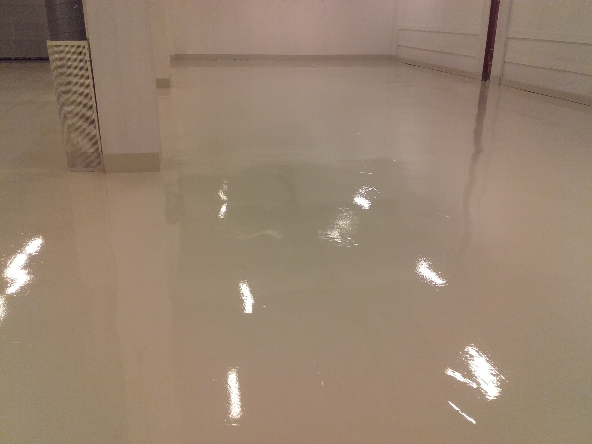 Błyszcząca, beżowa posadzka żywiczna w jasnym pomieszczeniu. Widoczne kolumny i fragmenty ścian. Gładka, refleksyjna powierzchnia podłogi.