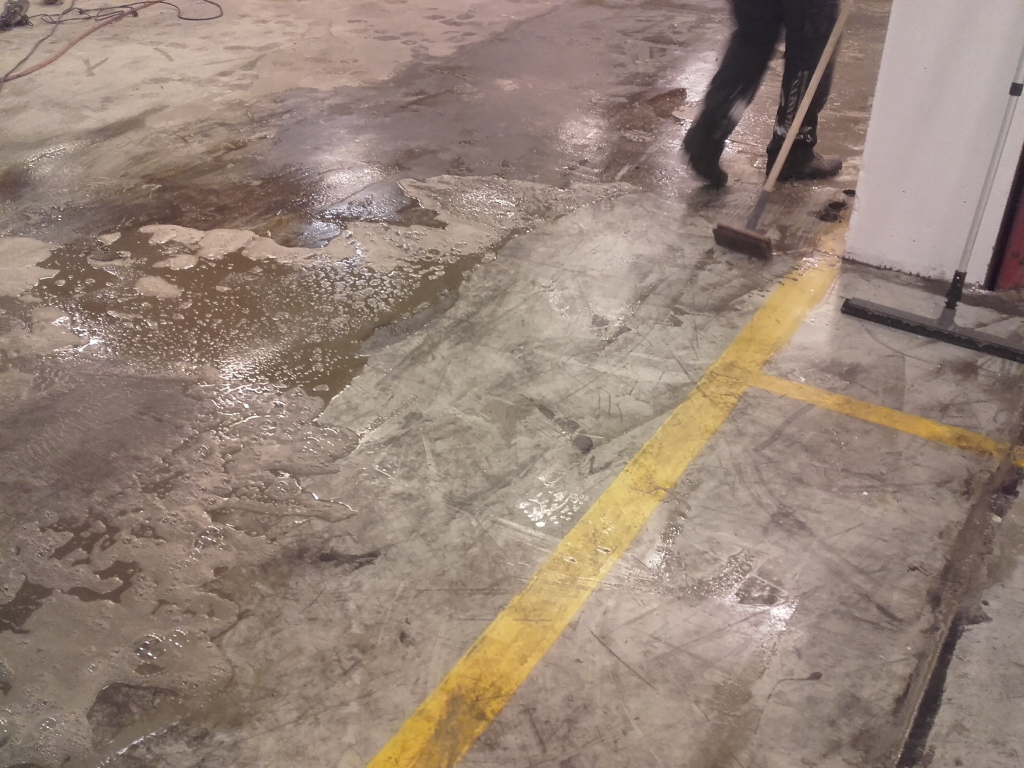 Czyszczenie posadzki przemysłowej z resztek wody i zabrudzeń za pomocą szczotki, widoczne żółte pasy na podłodze i osoba w butach roboczych.