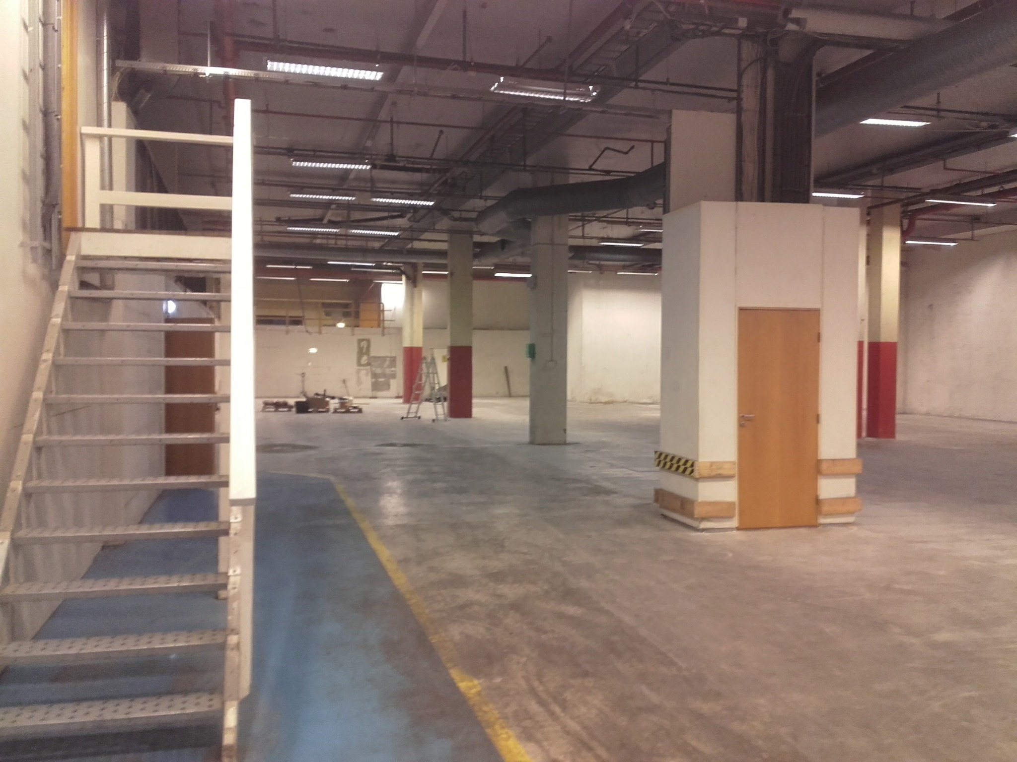 Przestronne wnętrze hali z metalowymi schodami po lewej, betonową podłogą z żółtą linią i filarami. Widoczne instalacje pod sufitem i drabina w tle.
