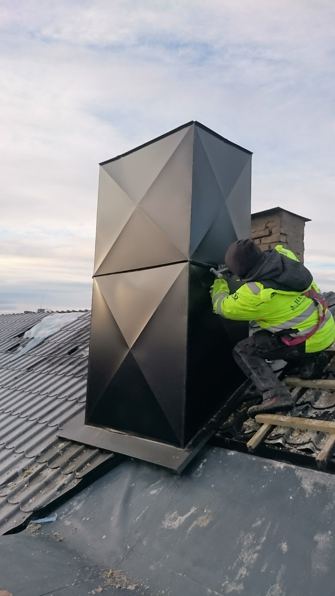 Pracownik w jaskrawej kurtce uszczelnia czarny, geometryczny komin na dachu z szarej dachówki, używając pistoletu do uszczelniacza. Widoczne drabiny dekarskie i fragment nieba.