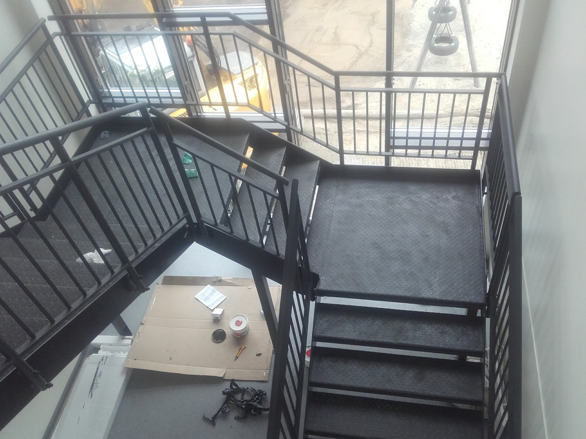 Metalowe schody z barierkami w trakcie malowania, widoczne narzędzia i puszka z farbą. Ujęcie z góry na klatkę schodową w budynku komercyjnym.