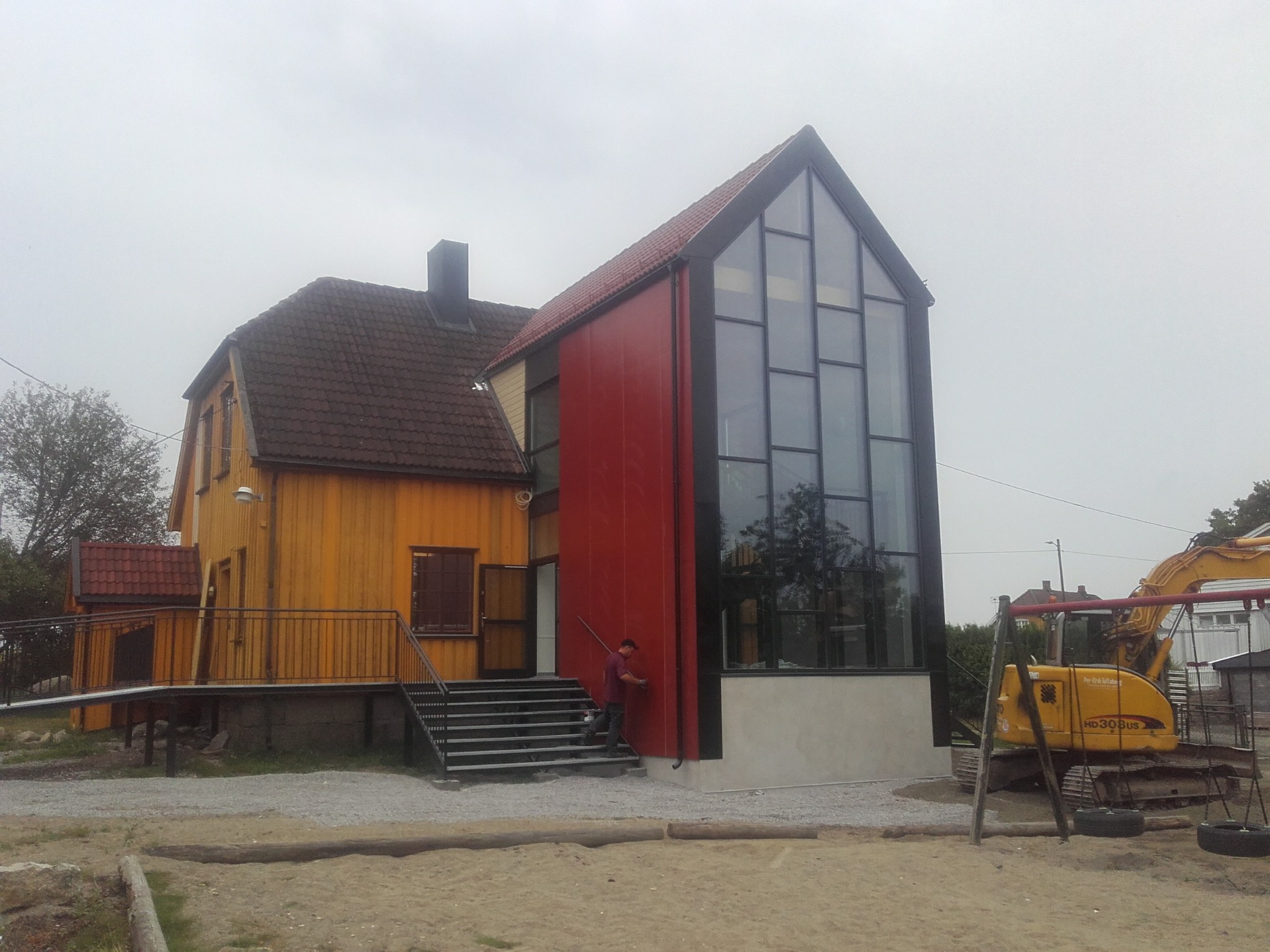 Żółty dom z czerwonym dachem i nowoczesną, przeszkloną dobudówką. Na pierwszym planie piasek i częściowo widoczna koparka. Osoba na schodach przy budynku.