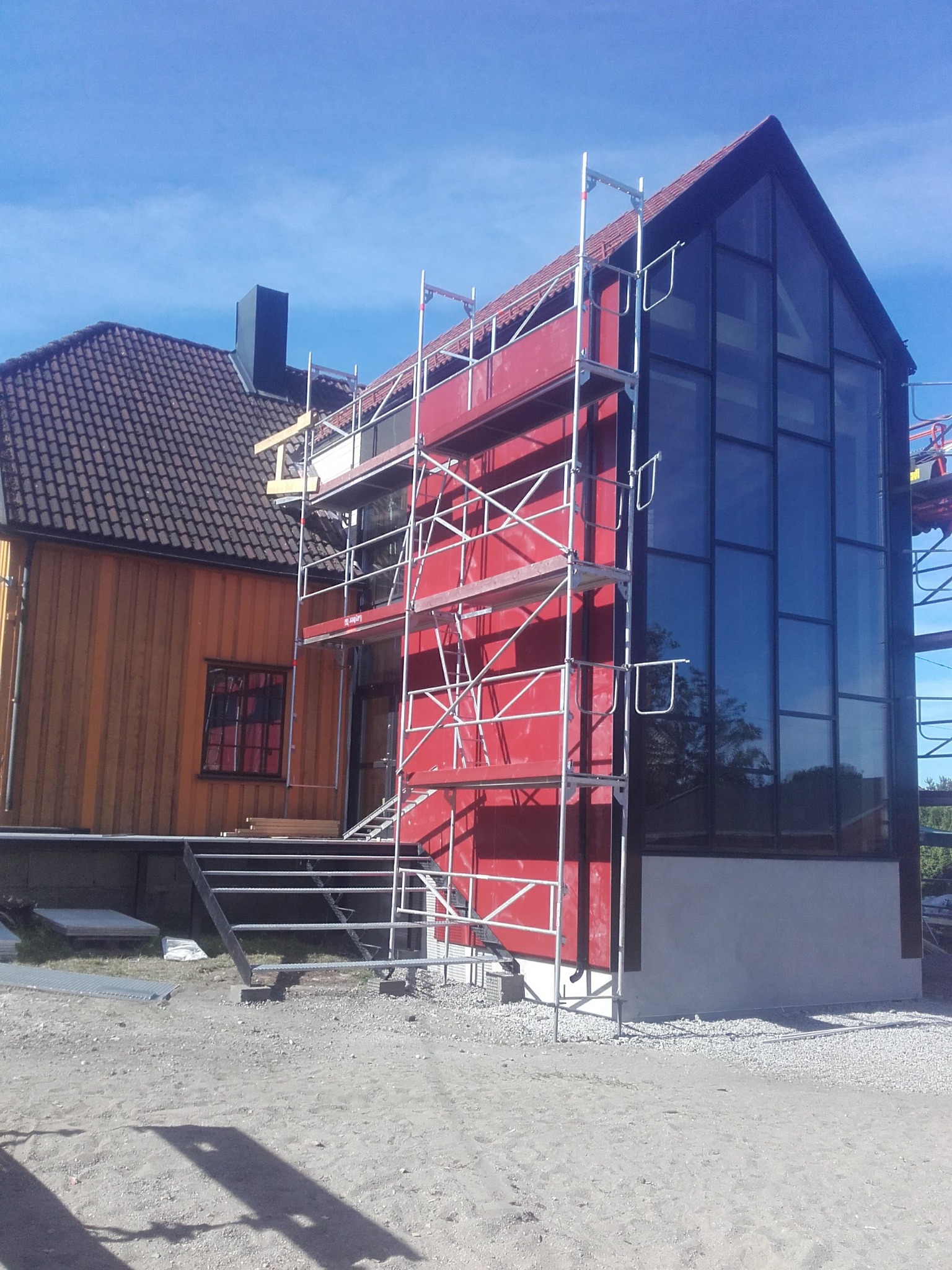 Dom w trakcie budowy z rusztowaniem, widoczne elementy konstrukcyjne i okna. Czerwona fasada kontrastuje z tradycyjną architekturą obok.