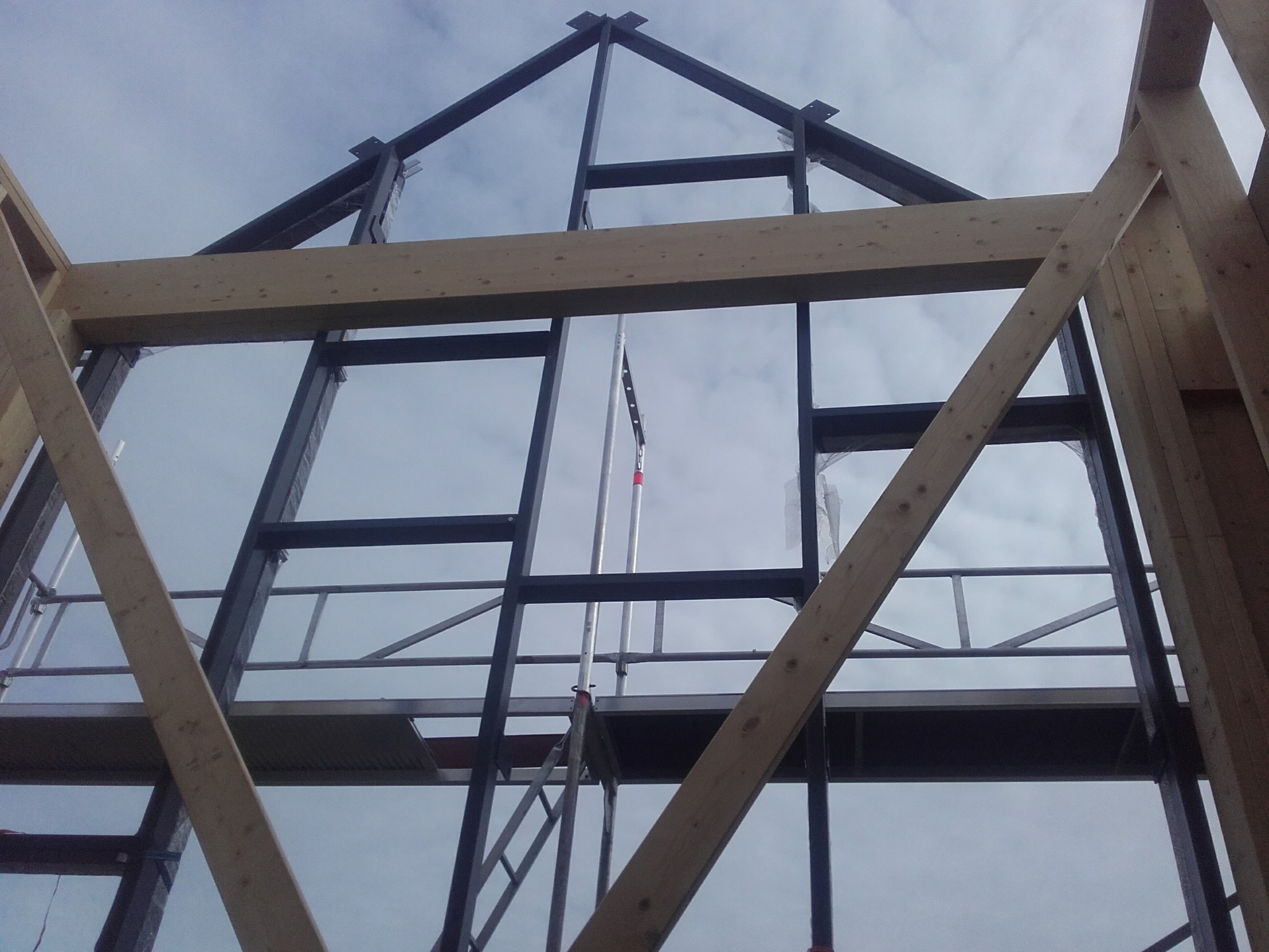 Wnętrze domu szkieletowego w budowie, widok z dołu na metalową konstrukcję dachu i drewniane elementy szkieletu na tle pochmurnego nieba.