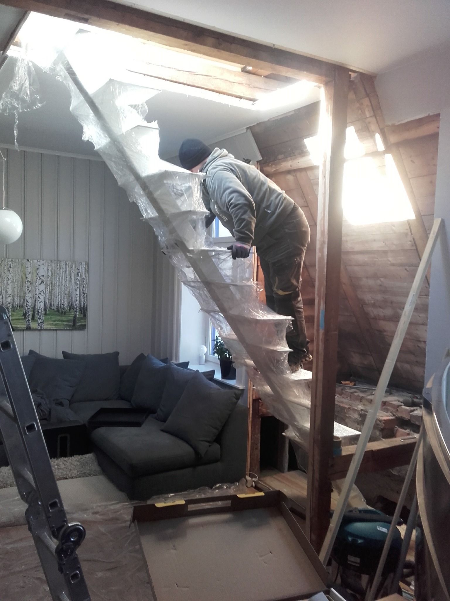 Pracownik w kombinezonie na schodach zabezpieczonych folią, w trakcie prac remontowych na poddaszu. Widoczne odsłonięte belki i cegły.