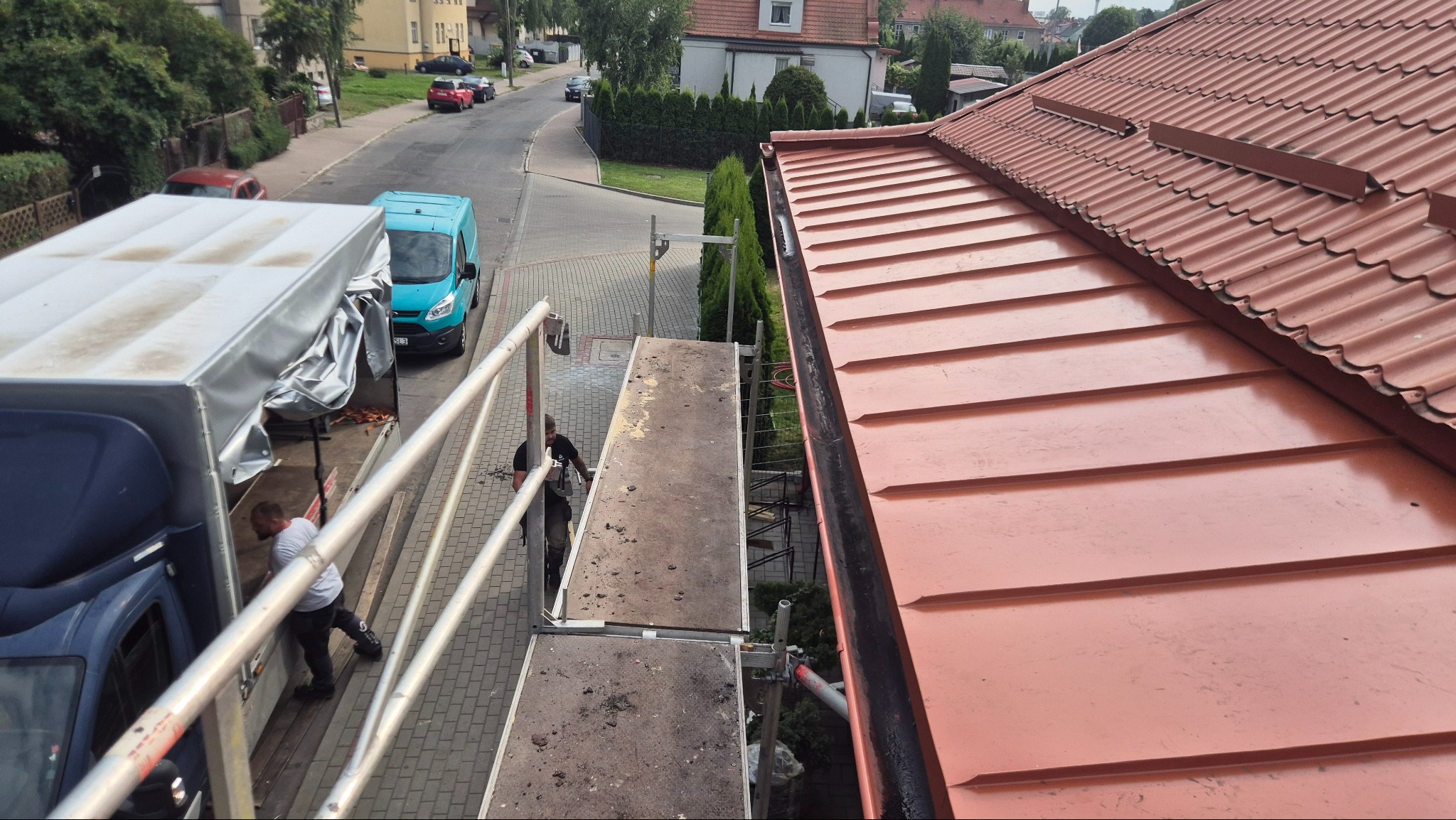 Czerwony dach z blachy na budynku, widok z rusztowania. Częściowo widoczny pracownik i samochód dostawczy. Ujęcie z góry, w tle zabudowa miejska.