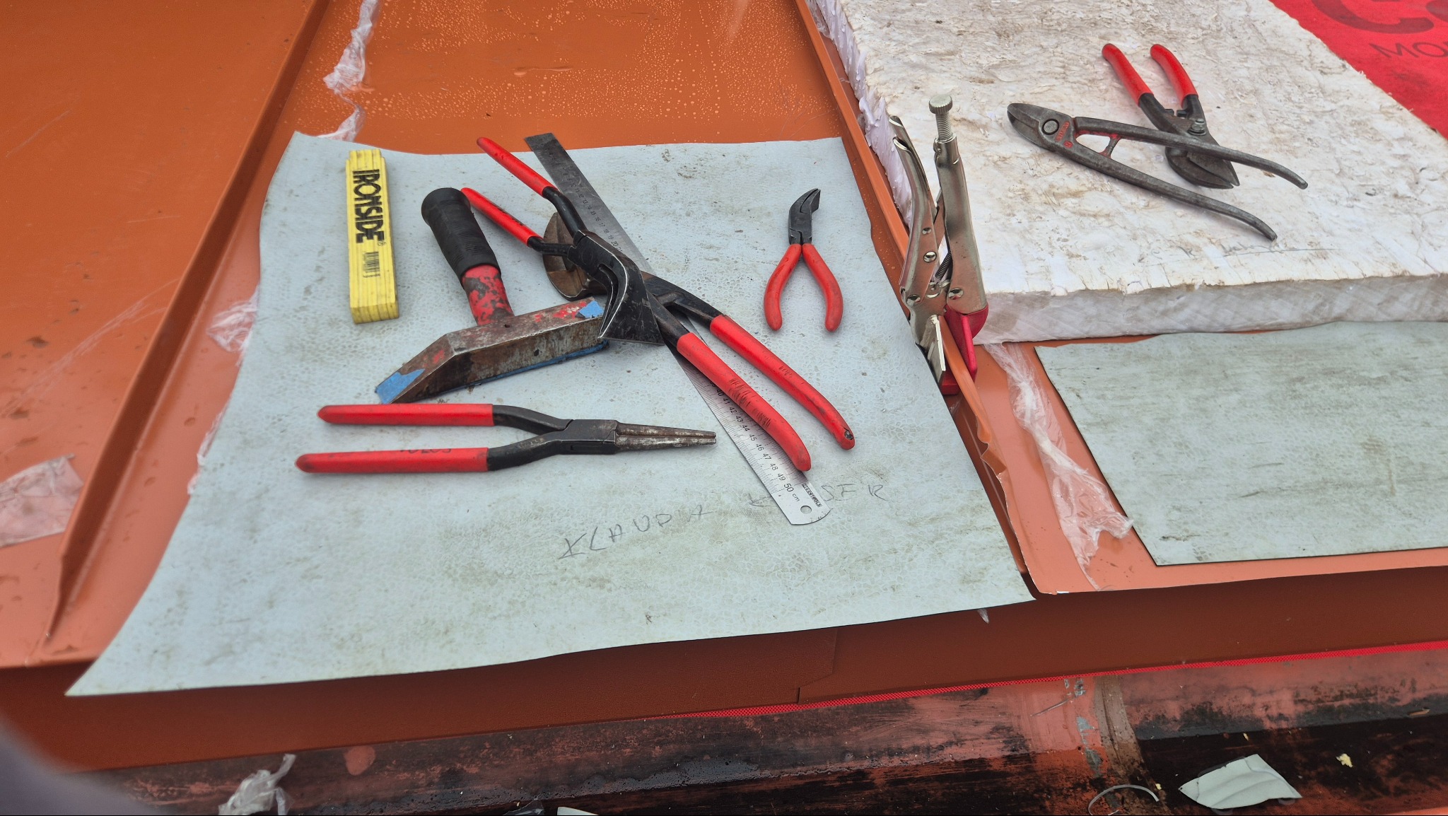 Narzędzia dekarskie, w tym młotek, kombinerki i miarka, ułożone na arkuszu blachy na dachu. Widoczne ocieplenie i elementy konstrukcji dachu.