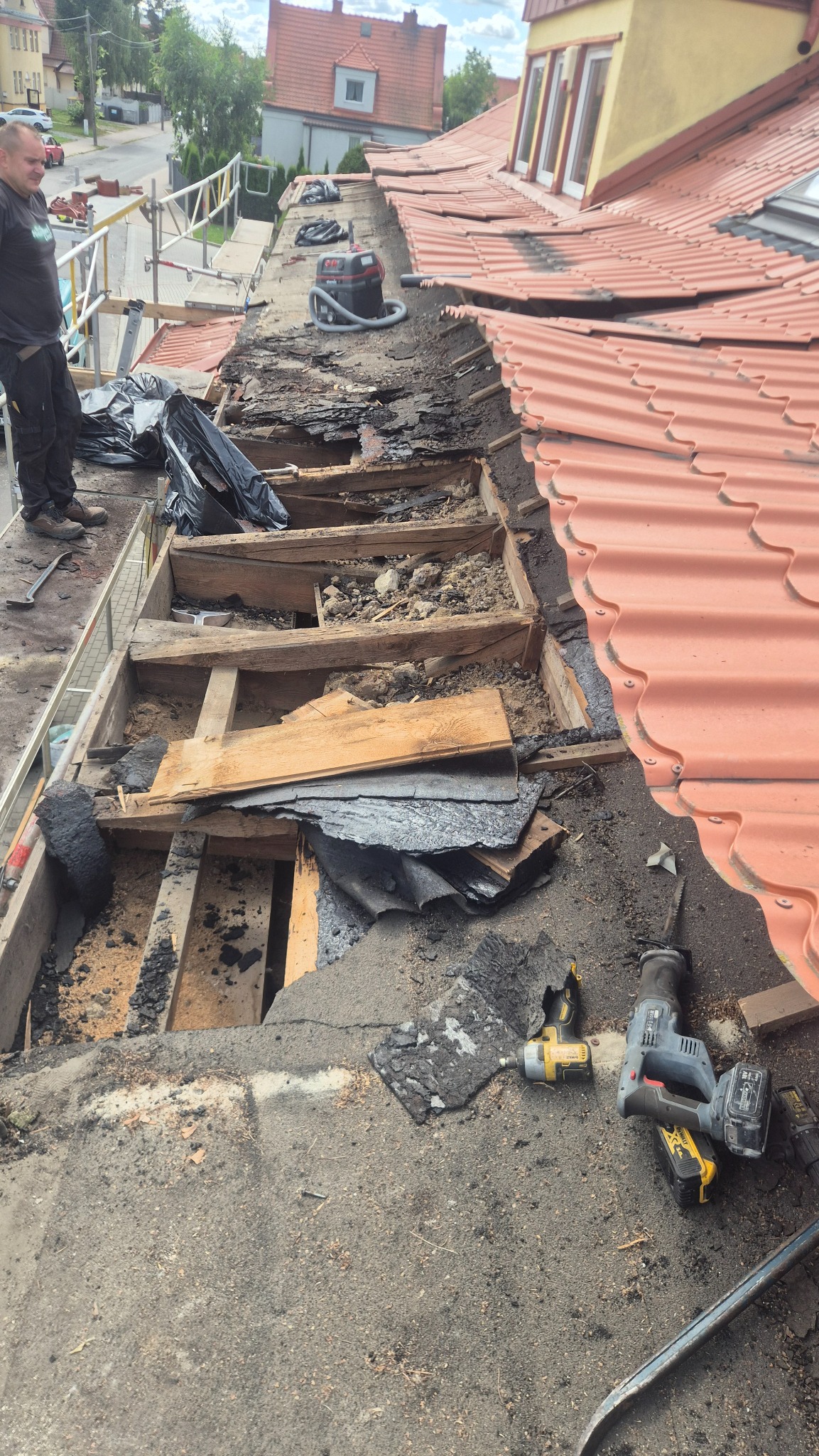 Demontaż starego dachu z odsłoniętą konstrukcją więźby, widoczne narzędzia i odkurzacz przemysłowy. Część dachu pokryta nową, czerwoną dachówką.