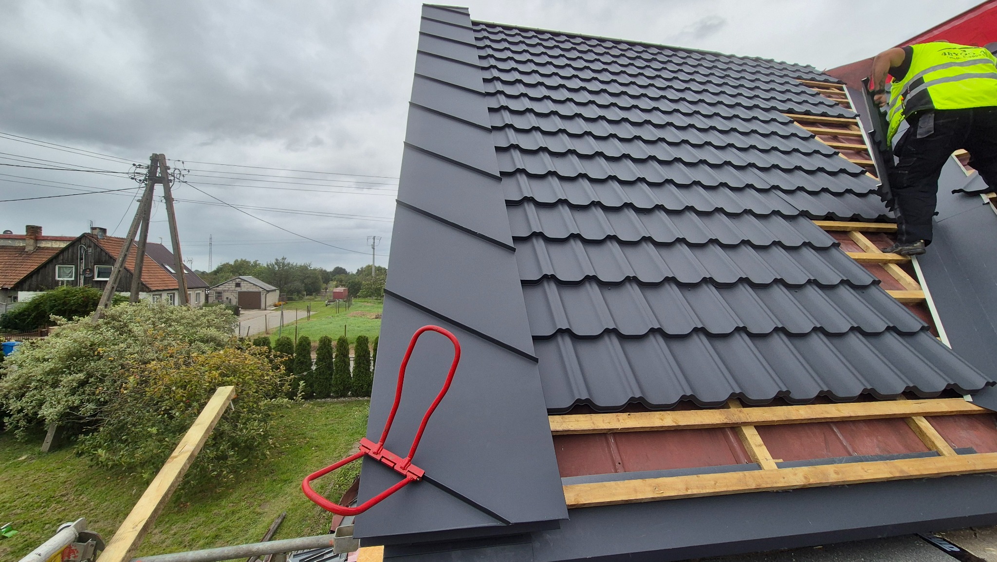 Montaż grafitowej blachodachówki na drewnianej konstrukcji dachu. Widoczny fragment pracownika w odblaskowej kamizelce i narzędzia dekarskie. Tło: zabudowa wiejska.