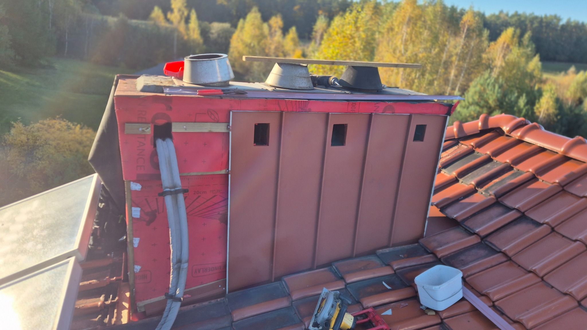 Wysoki komin w trakcie obróbki blacharskiej na dachu z brązowej dachówki. Widoczne narzędzia, elementy wentylacyjne i membrana dachowa. Tło stanowią drzewa.