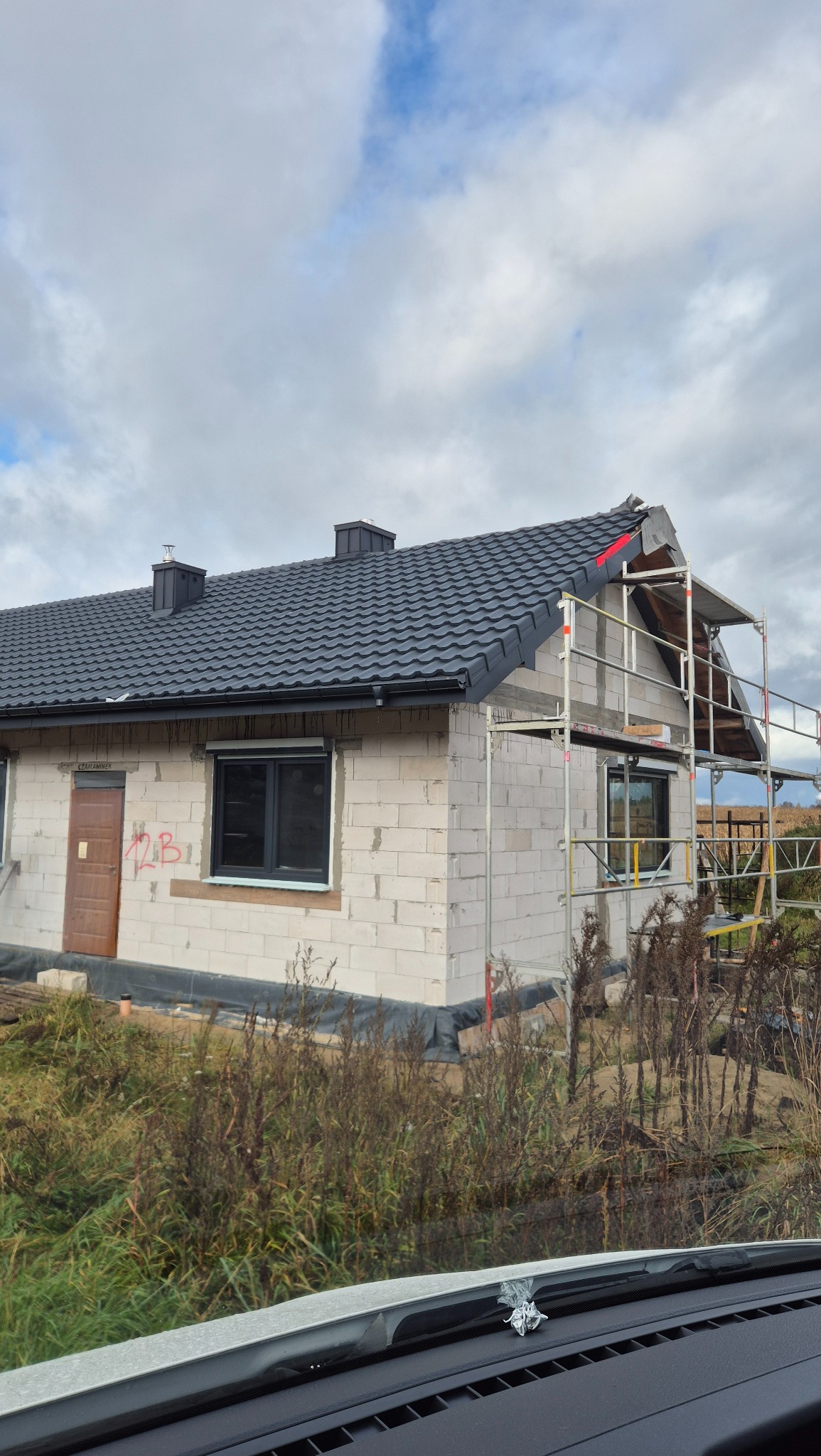 Dom w trakcie budowy z czarną dachówką i rusztowaniem. Widoczne bloki betonowe i elementy konstrukcyjne dachu. Częściowo ukończony dach z rynnami.