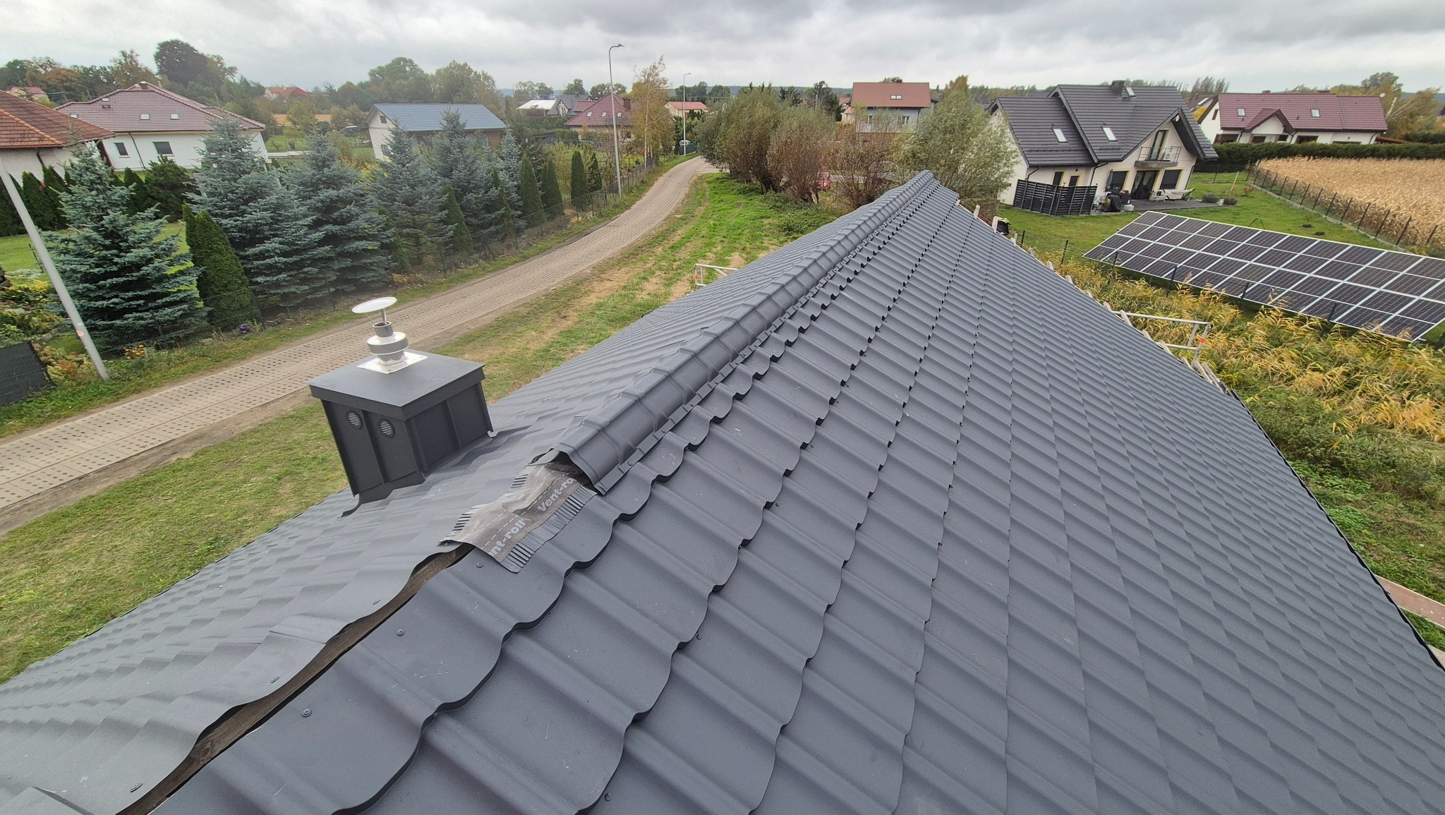 Szary dach z blachy trapezowej, komin z czarną obudową. W tle domy jednorodzinne, panele słoneczne i droga. Widok z góry na nowo pokryty dach.