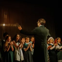 Chór młodzieżowy na scenie z dyrygentem w fraku, w trakcie występu, z rękami złożonymi do oklasków, w ciemnym oświetleniu.