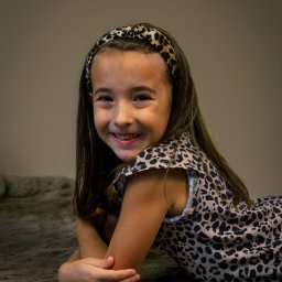 ONE SPÓŁKA Z OGRANICZONĄ ODPOWIEDZIALNOŚCIĄ - Portret uśmiechniętej dziewczynki w stroju w panterkę, leżącej na futrzanym dywanie z założonymi rękami, z opaską na włosach w tym samym wzorze.