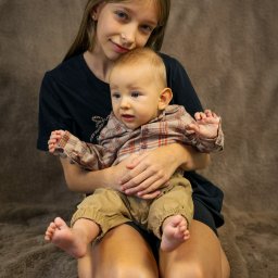 ONE SPÓŁKA Z OGRANICZONĄ ODPOWIEDZIALNOŚCIĄ - Portret dziewczynki trzymającej na kolanach małego chłopca w koszuli w kratę, na tle brązowego koca z długim włosiem. Dziecko patrzy w bok.