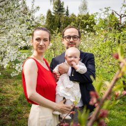 ONE SPÓŁKA Z OGRANICZONĄ ODPOWIEDZIALNOŚCIĄ - Rodzinne zdjęcie w plenerze: mama w czerwonej bluzce i tata w marynarce trzymają ubraną w białą sukienkę córeczkę na tle kwitnących drzew.