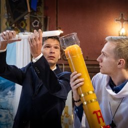 ONE SPÓŁKA Z OGRANICZONĄ ODPOWIEDZIALNOŚCIĄ - Uroczystość w kościele: mężczyzna w garniturze zapala świecę od paschału trzymanego przez ministranta. W tle ołtarz i inni uczestnicy ceremonii.