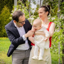 ONE SPÓŁKA Z OGRANICZONĄ ODPOWIEDZIALNOŚCIĄ - Uśmiechnięta rodzina z niemowlęciem w kwitnącym sadzie. Dziecko w białej sukience, rodzice w eleganckich strojach. Naturalne światło, wiosenna sceneria.