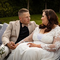 ONE SPÓŁKA Z OGRANICZONĄ ODPOWIEDZIALNOŚCIĄ - Para młoda siedzi na sofie, trzymając się za ręce. Pan młody w beżowym garniturze i muszce, panna młoda w białej sukni z koronkowymi rękawami. W tle zieleń.