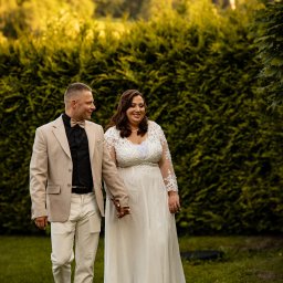 ONE SPÓŁKA Z OGRANICZONĄ ODPOWIEDZIALNOŚCIĄ - Uśmiechnięta para młoda, trzymając się za ręce, spaceruje po zielonym trawniku na tle wysokiego, zielonego żywopłotu w słoneczny dzień.