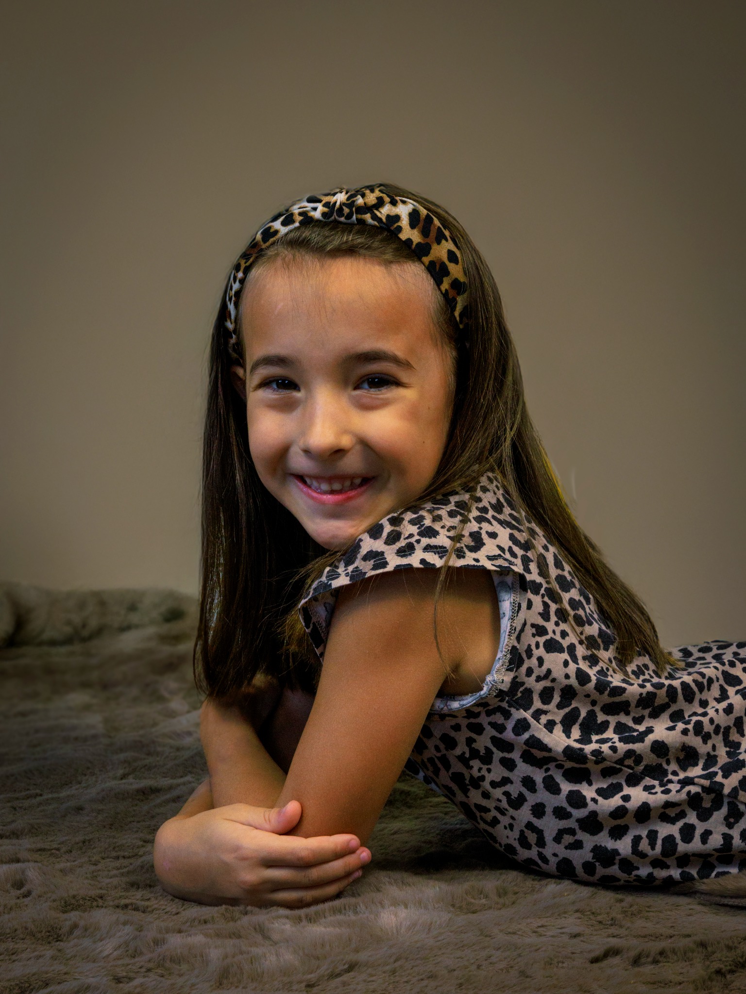 Portret uśmiechniętej dziewczynki w stroju w panterkę, leżącej na futrzanym dywanie z założonymi rękami, z opaską na włosach w tym samym wzorze.