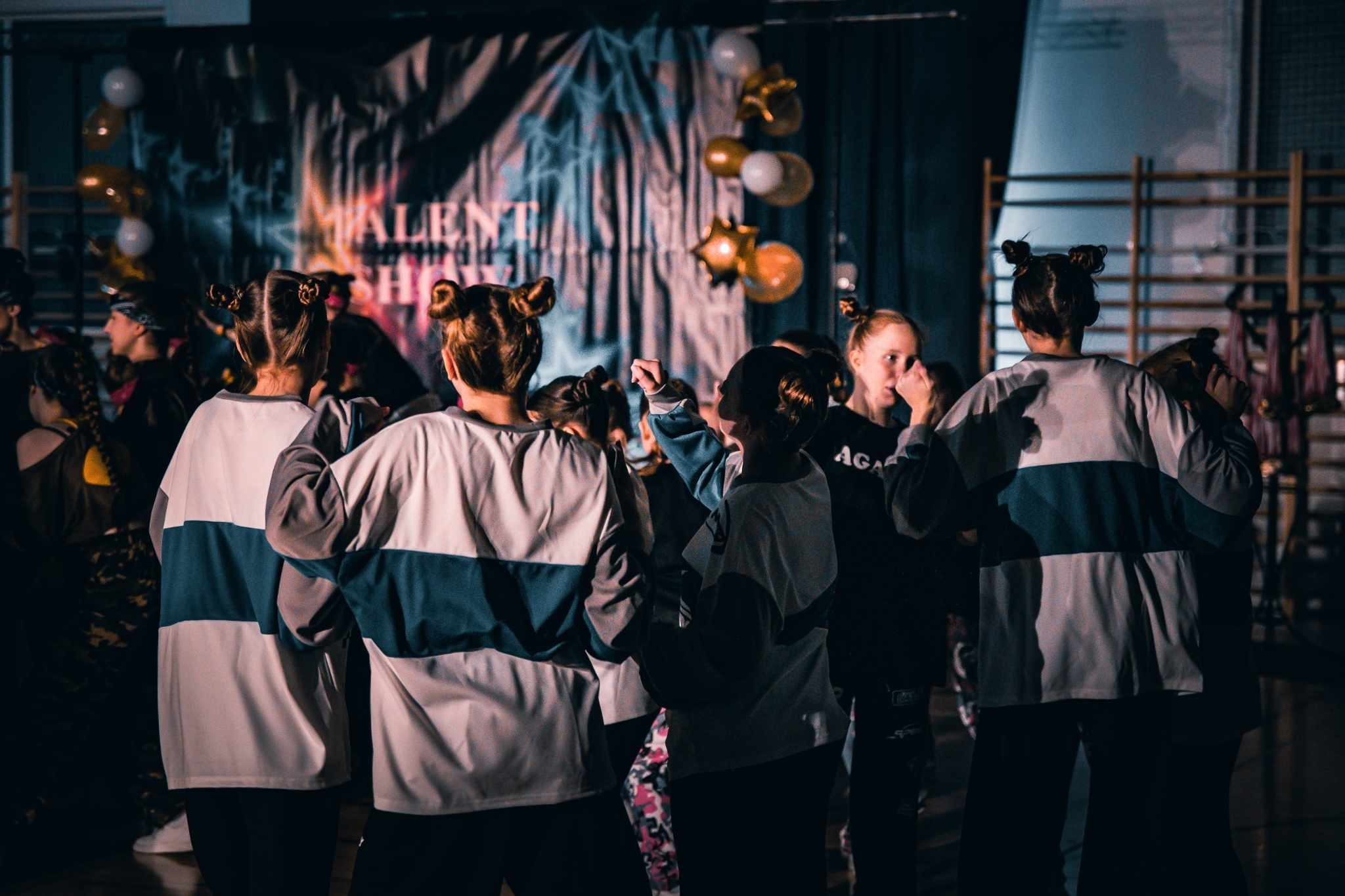 Grupa młodych tancerzy w sportowych strojach, z fryzurami w koczki, stoi tyłem do widza na tle sceny z napisem 'Talent Show' i balonami.