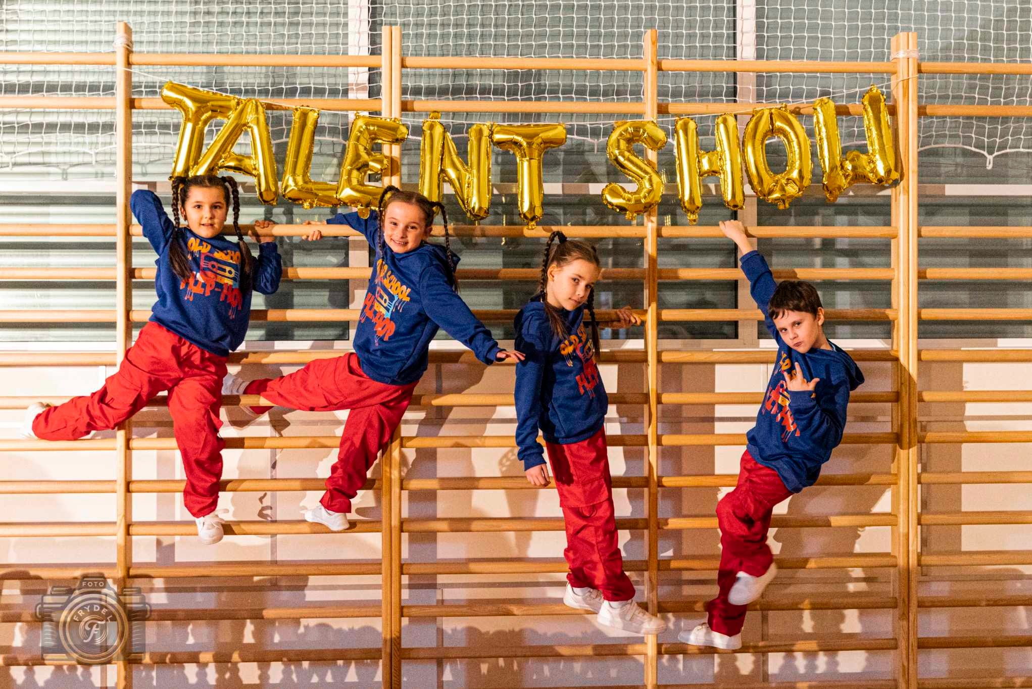 Czwórka dzieci w czerwonych spodniach i niebieskich bluzach pozuje na drabinkach w sali gimnastycznej pod złotym napisem 'TALENT SHOW'.