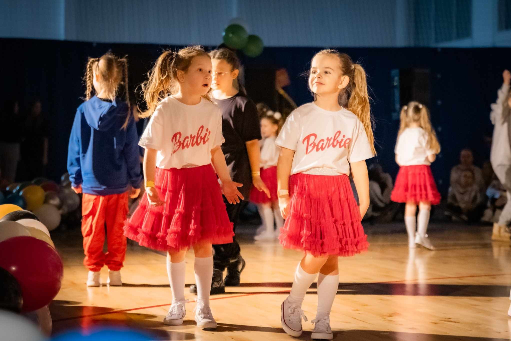 Grupa dzieci w kostiumach 'Barbie' na scenie podczas występu. Dwie dziewczynki na pierwszym planie, w białych koszulkach z napisem 'Barbie' i czerwonych tiulowych spódniczkach.