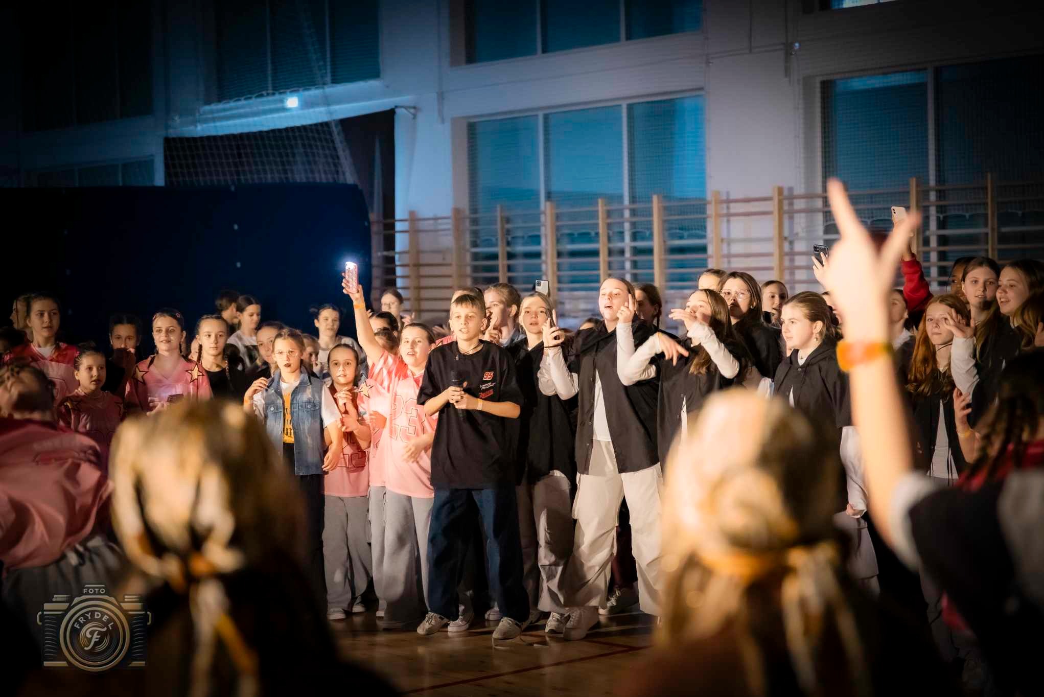 Grupa młodzieży w sali gimnastycznej, część z telefonami w dłoniach, oświetlona reflektorami, prawdopodobnie podczas występu lub próby. W tle drabinki gimnastyczne.